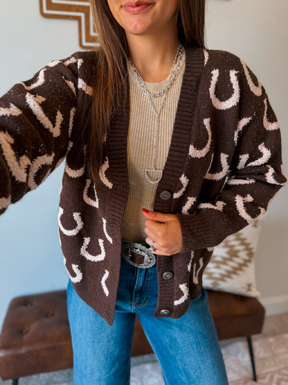 A woman wearing a brown knit cardigan with a cream horseshoe pattern, layered over a beige ribbed top and blue jeans.