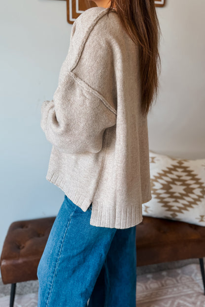 A woman wearing a chunky knit beige sweater and blue jeans, shown from a side-back angle in an indoor setting with a patterned pillow and brown bench in the background.