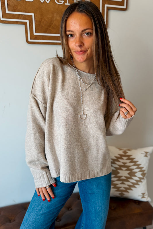 A woman wearing a light beige knit crewneck sweater and blue jeans, standing in an indoor setting with a patterned pillow and wooden wall decor in the background.