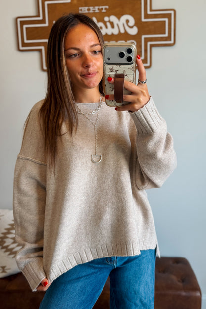 A woman takes a mirror selfie wearing a light beige knit sweater and blue jeans in a home setting.