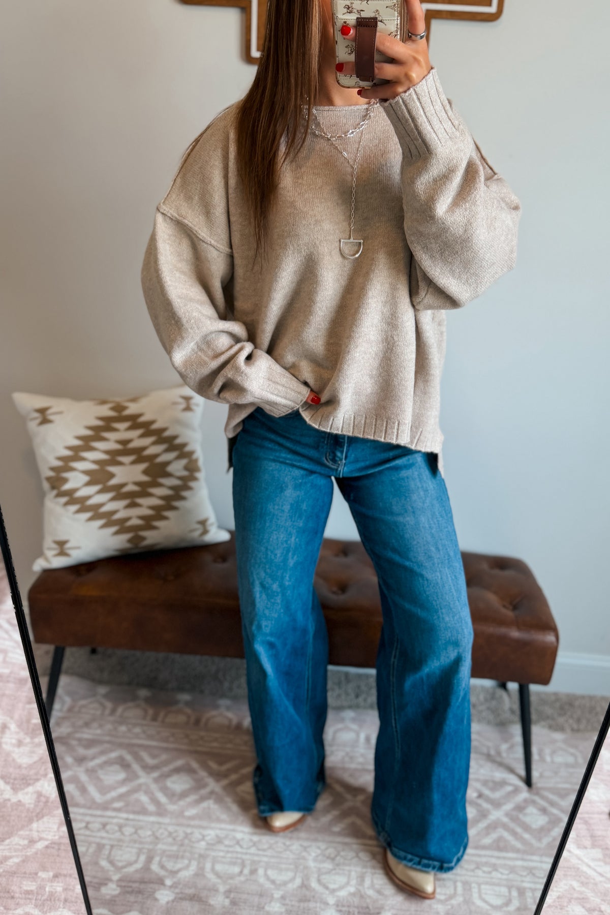 A woman takes a mirror selfie showing an outfit consisting of a beige knit crewneck sweater, blue wide-leg jeans, and beige pointed-toe flats. She is standing in a room with a brown leather tufted bench and a patterned throw pillow.