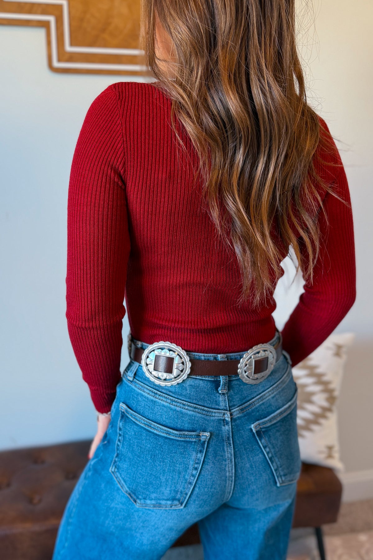 A woman shown from behind wearing a fitted red ribbed long-sleeve top, blue jeans, and a black belt with large silver concho-style buckles.