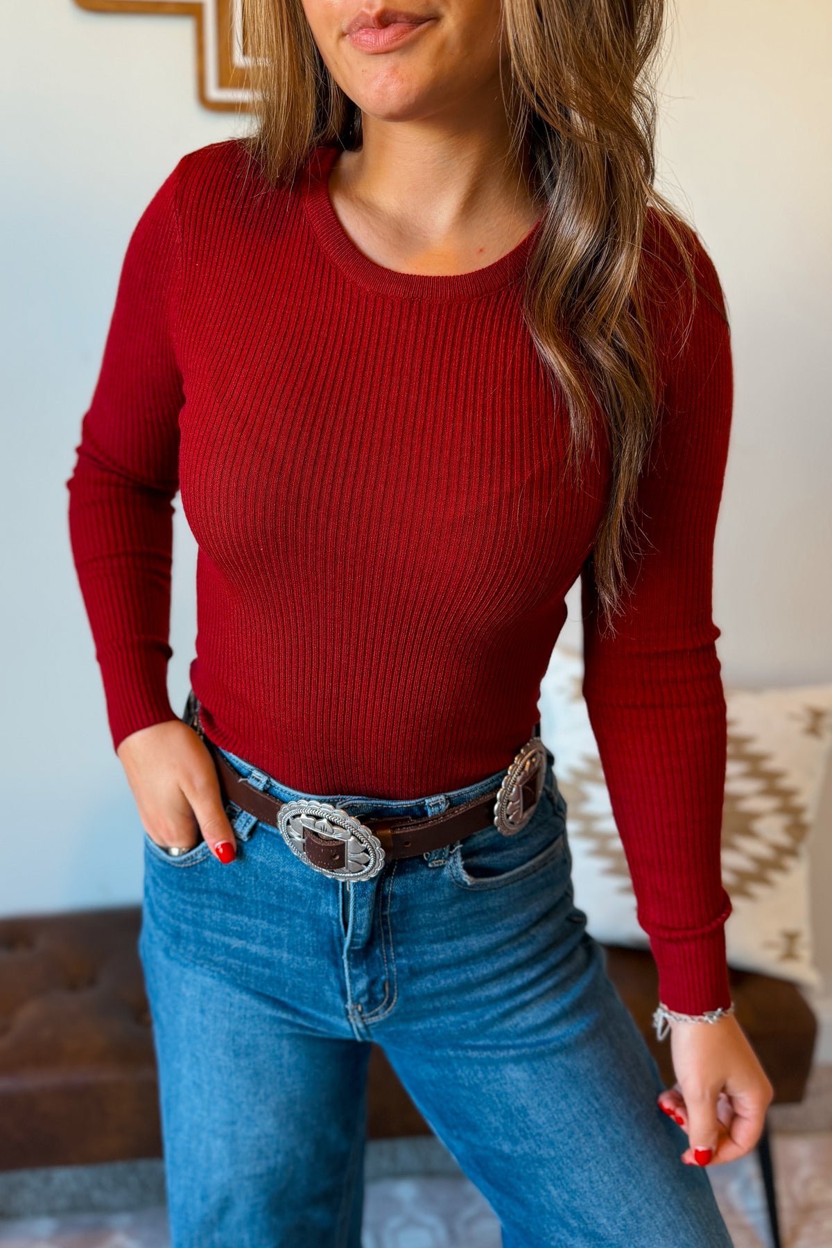 A woman wearing a fitted long-sleeve ribbed knit sweater in deep red, paired with blue jeans and a western-style belt, shown from the chest down in an indoor setting.