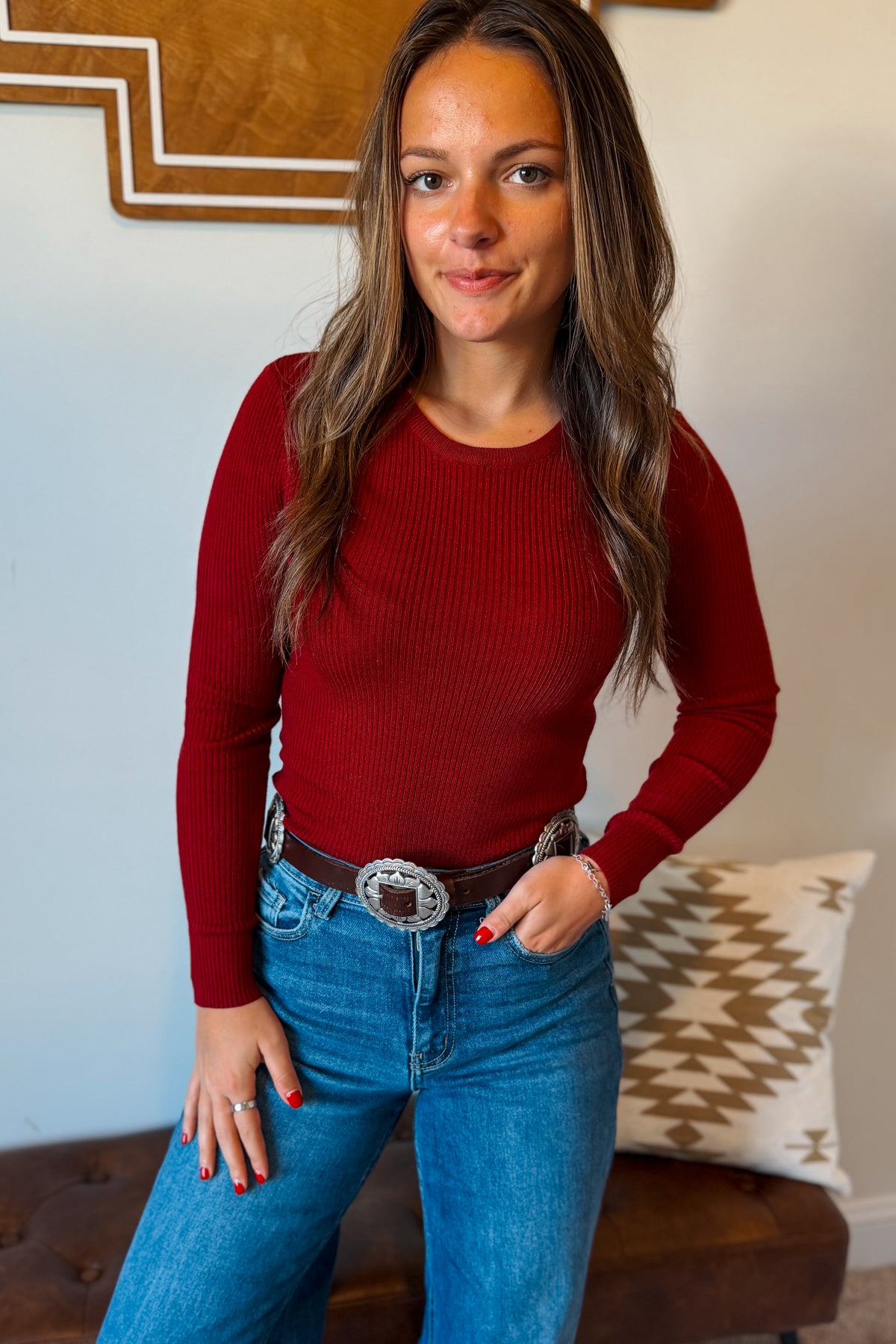 A woman wearing a fitted long-sleeve ribbed knit top in deep red, paired with blue jeans and a brown belt with a large silver buckle, standing indoors.