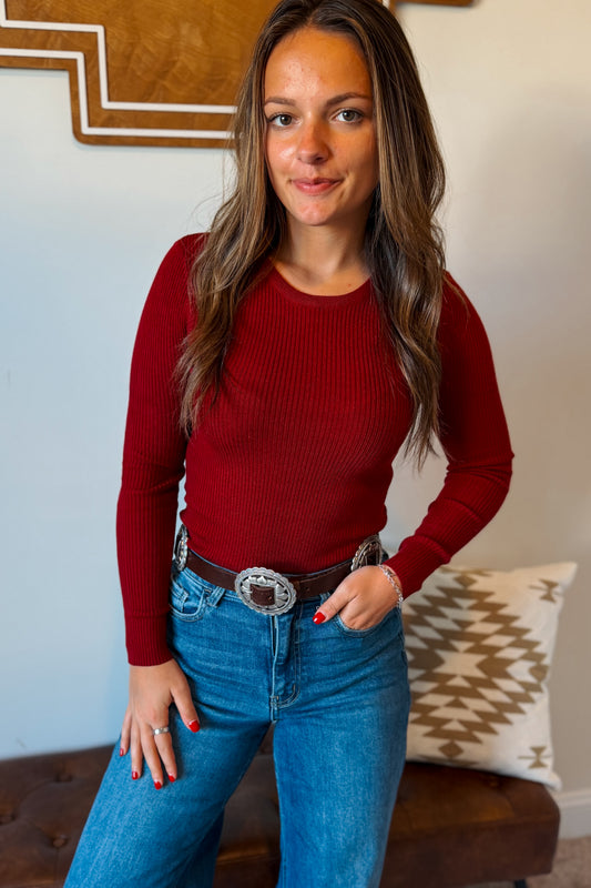 A woman wearing a fitted long-sleeve ribbed knit top in deep red, paired with blue jeans and a brown belt with a large silver buckle, standing indoors.