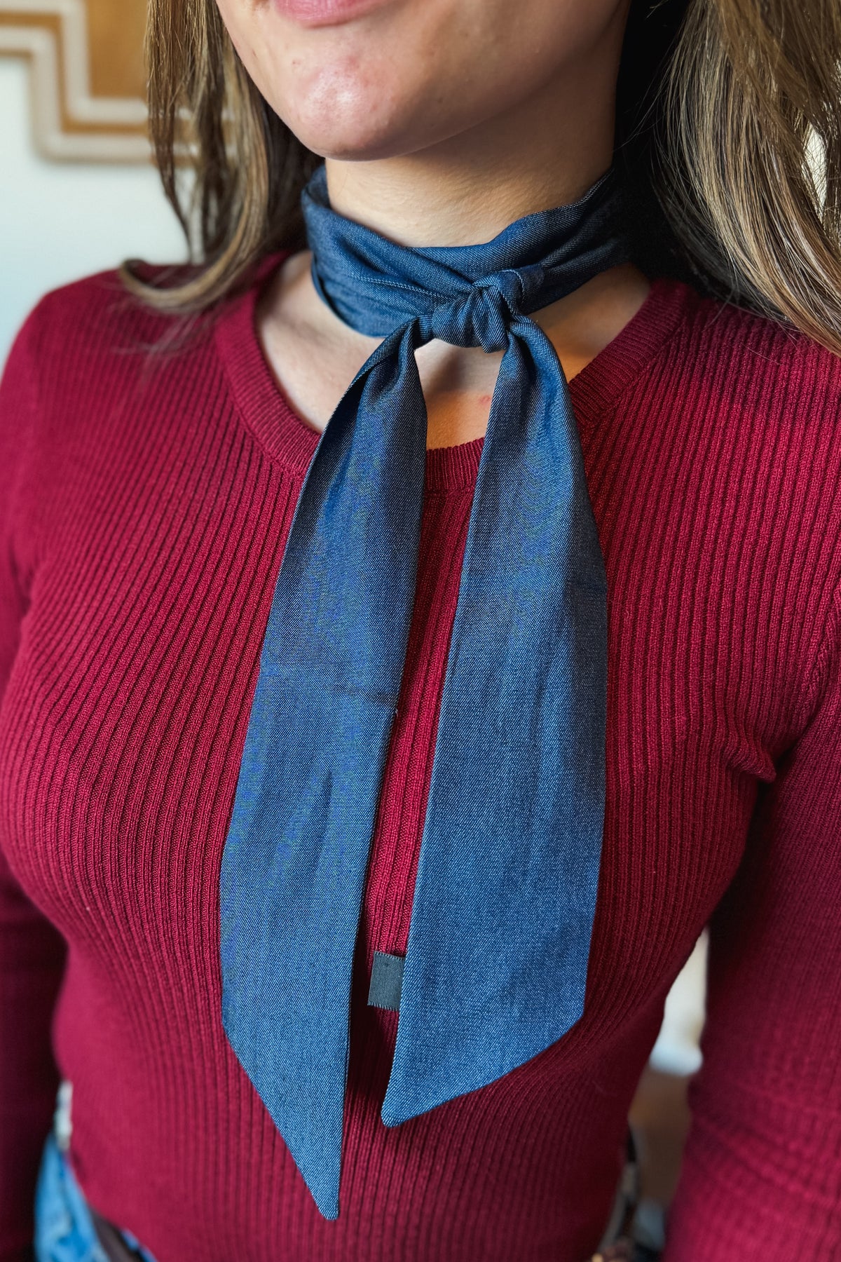 A close-up of a person wearing a slate blue linen neck scarf tied in a knot over a ribbed burgundy sweater.