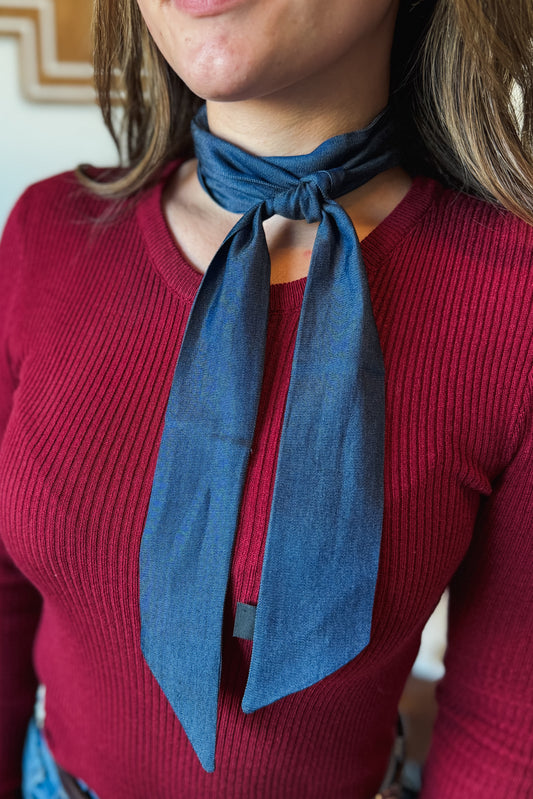 A close-up of a person wearing a slate blue linen neck scarf tied in a knot over a ribbed burgundy sweater.