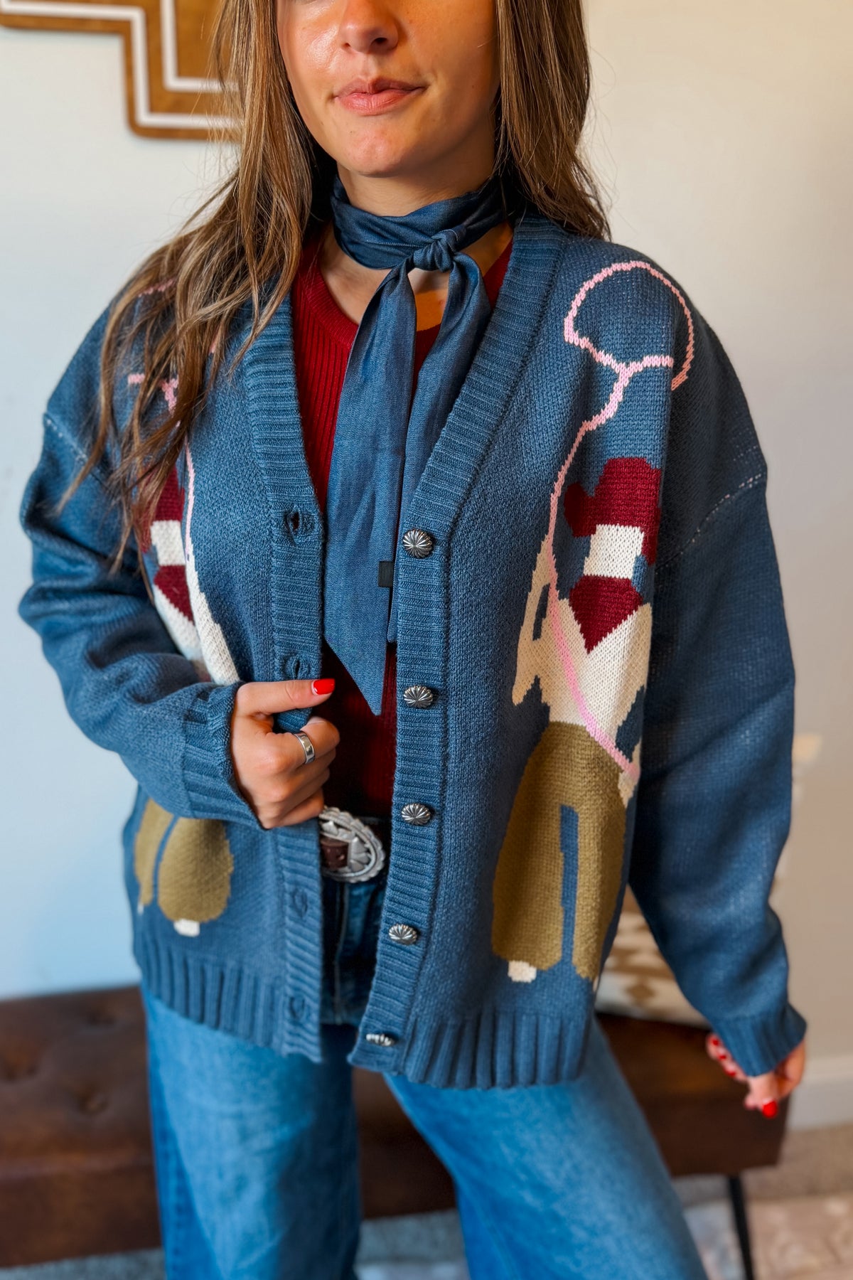 A woman wearing a blue knit cardigan with a western-themed donkey pattern in red, beige, and white, layered over a red top and blue jeans.