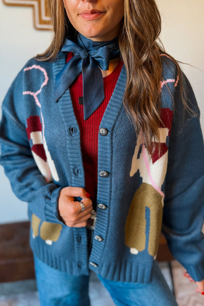 A woman wearing a blue knit cardigan with a colorful geometric pattern, a dark blue necktie, and a red ribbed top, paired with blue jeans.