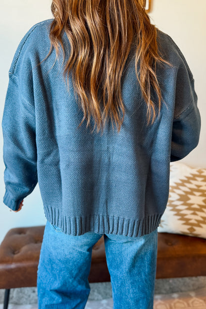 A person viewed from behind wearing an oversized dusty blue knit sweater and blue jeans, standing indoors near a brown bench and a patterned pillow.