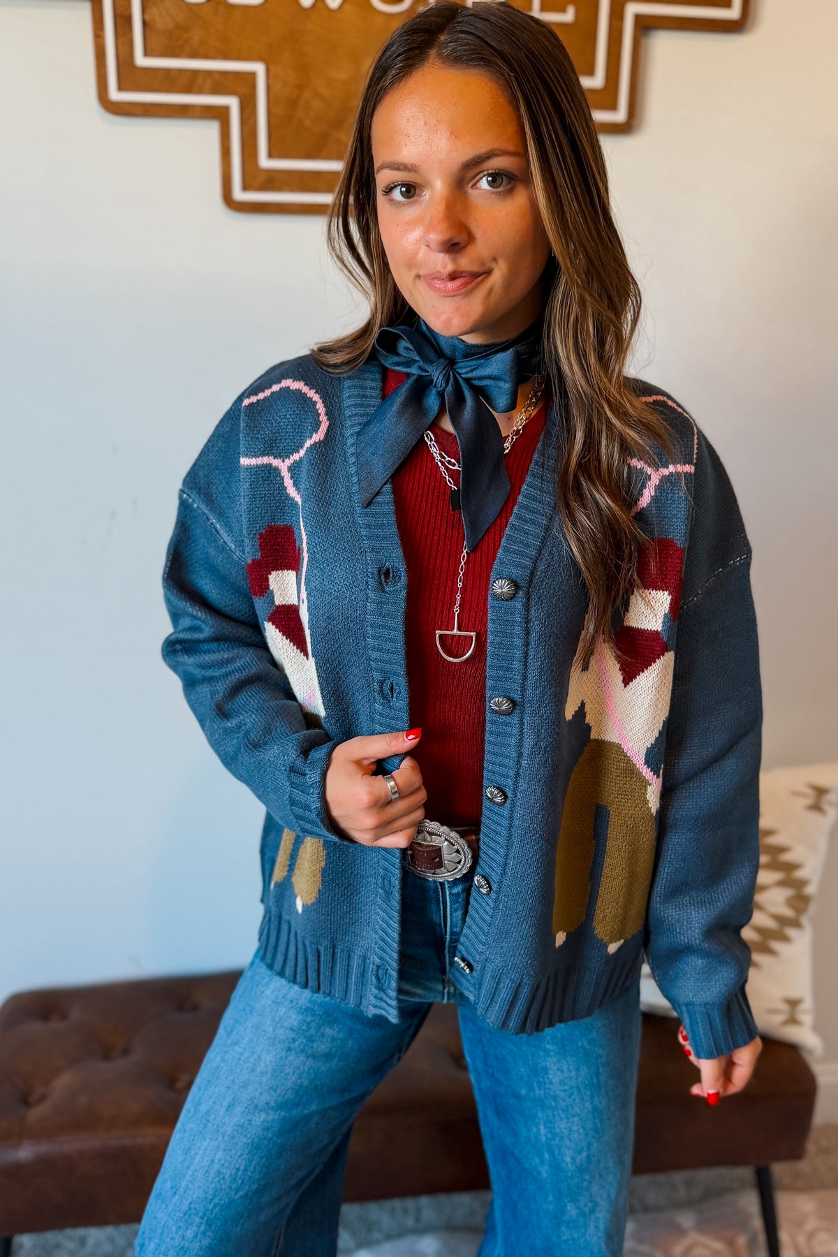 A woman wearing a blue knit cardigan with western-style geometric and animal motifs, layered over a red sweater, blue jeans, and a blue necktie, standing in an indoor setting.
