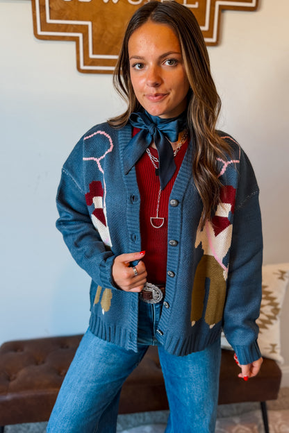 A woman wearing a blue knit cardigan with western-style geometric and animal motifs, layered over a red sweater, blue jeans, and a blue necktie, standing in an indoor setting.