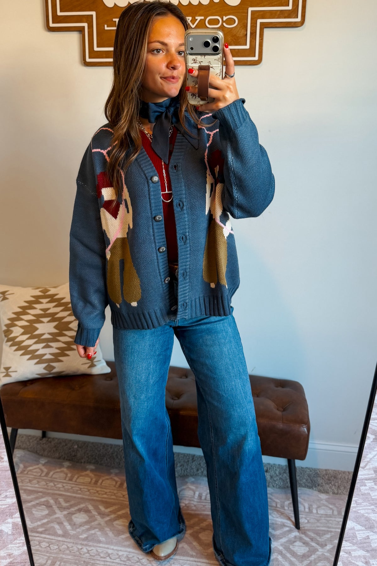 A woman takes a mirror selfie wearing a blue knit cardigan with a colorful geometric pattern and wide-leg blue jeans, standing in a room with a brown tufted bench and a patterned rug.