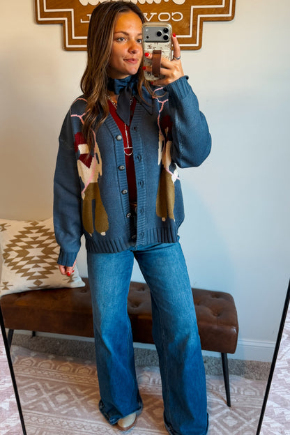 A woman takes a mirror selfie wearing a blue knit cardigan with a colorful geometric pattern and wide-leg blue jeans, standing in a room with a brown tufted bench and a patterned rug.