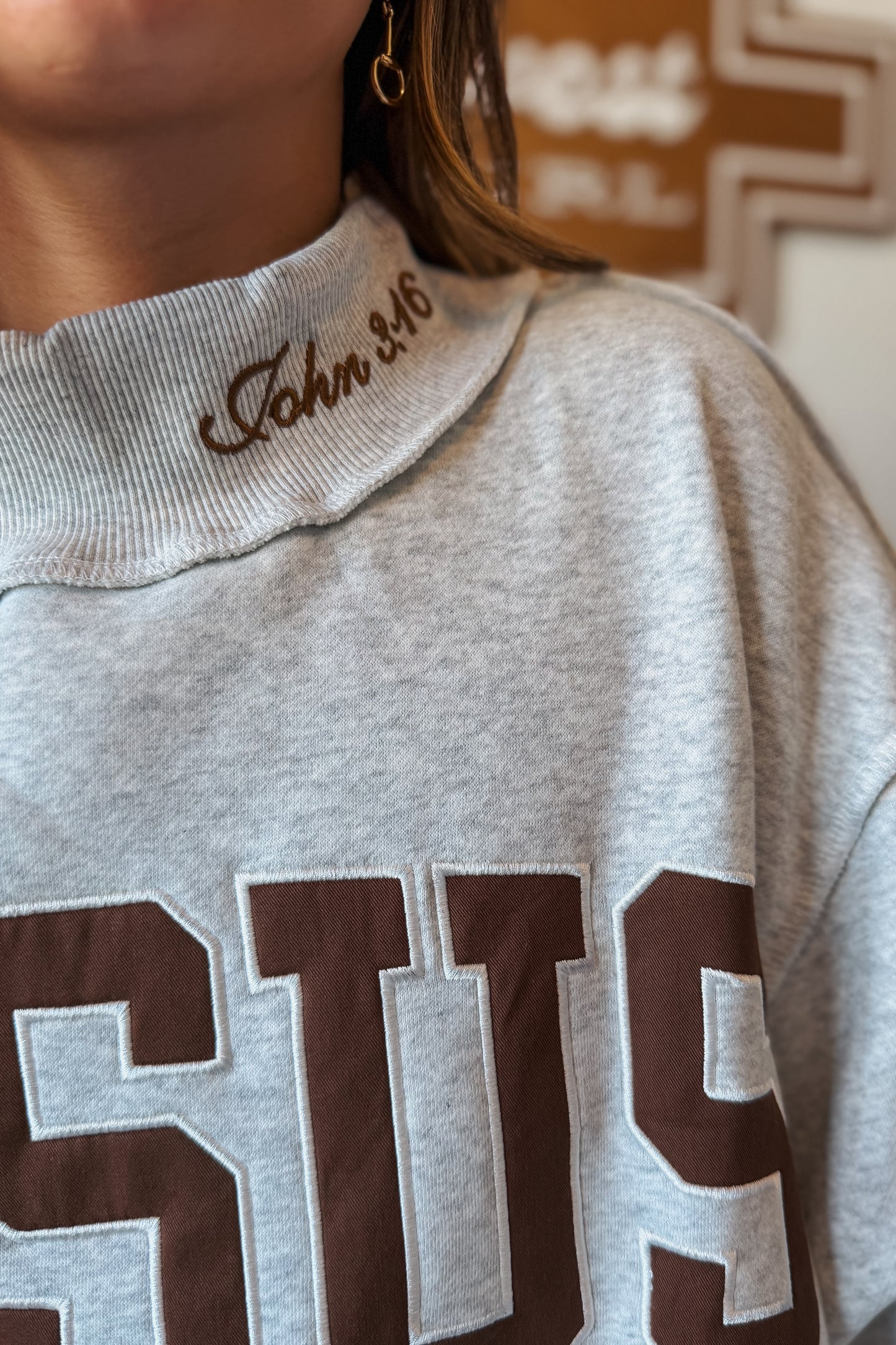 Close-up view of a brown mock neck sweatshirt highlighting the embroidered "Jesus" lettering and John 3:16 scripture detail stitched along the neckline.