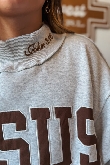 Close-up view of a brown mock neck sweatshirt highlighting the embroidered "Jesus" lettering and John 3:16 scripture detail stitched along the neckline.