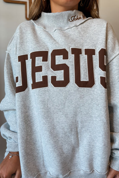Close-up view of a brown mock neck sweatshirt highlighting the embroidered "Jesus" lettering and John 3:16 scripture detail stitched along the neckline.