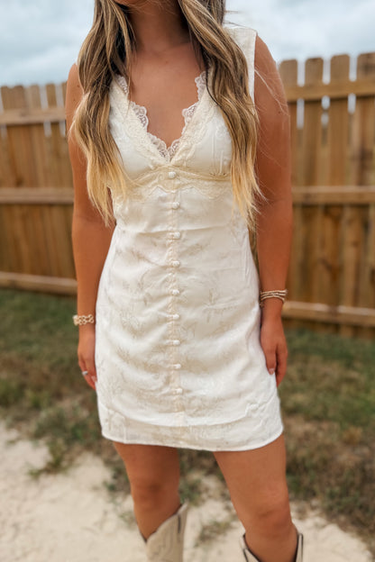 A woman wearing a sleeveless cream-colored lace and textured mini dress with a button-down front, standing outdoors in front of a wooden fence.