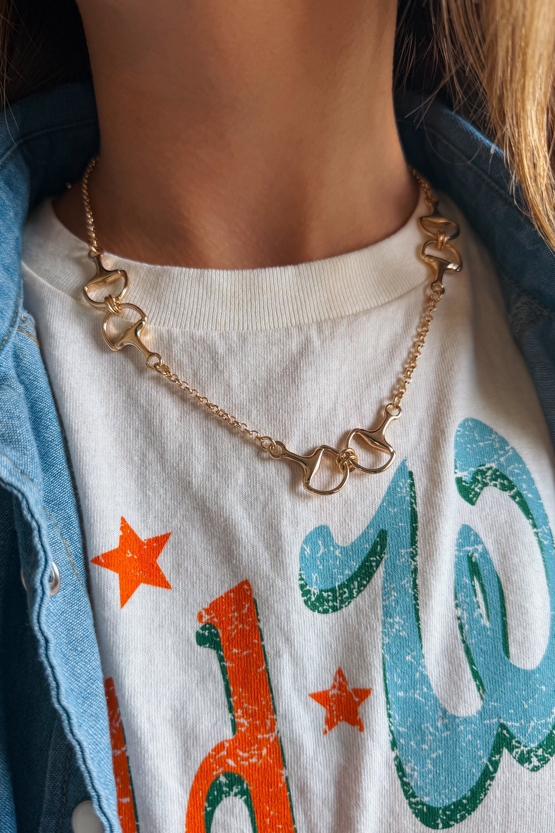 A close-up of a person wearing a gold horse-bit chain necklace over a white graphic t-shirt and a light blue denim jacket.