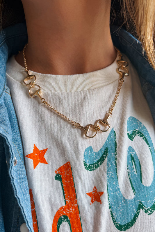 A close-up of a person wearing a gold horse-bit chain necklace over a white graphic t-shirt and a light blue denim jacket.