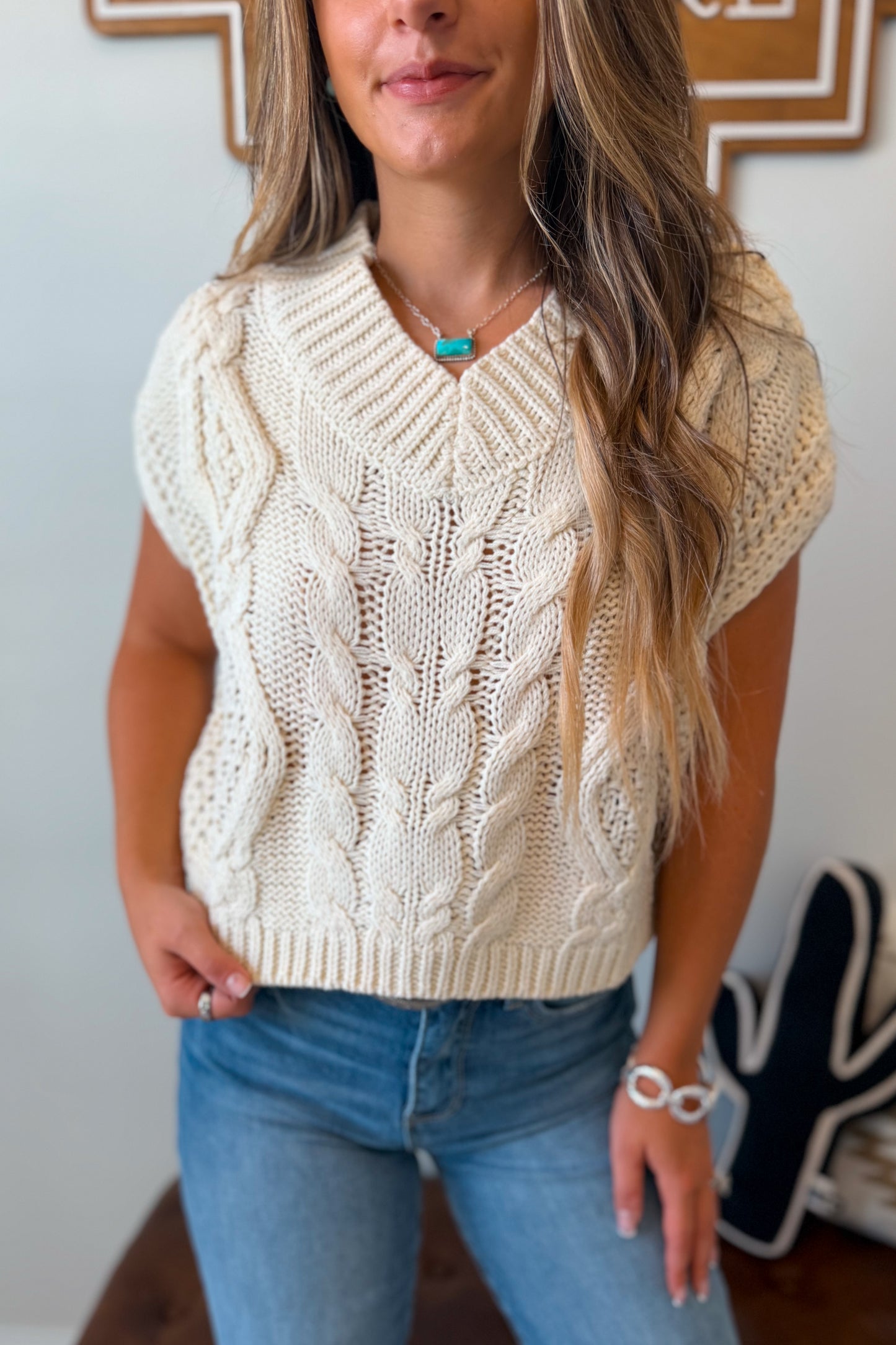 Close Up of A woman wearing a cream cable-knit short-sleeve V-neck sweater vest paired with blue jeans