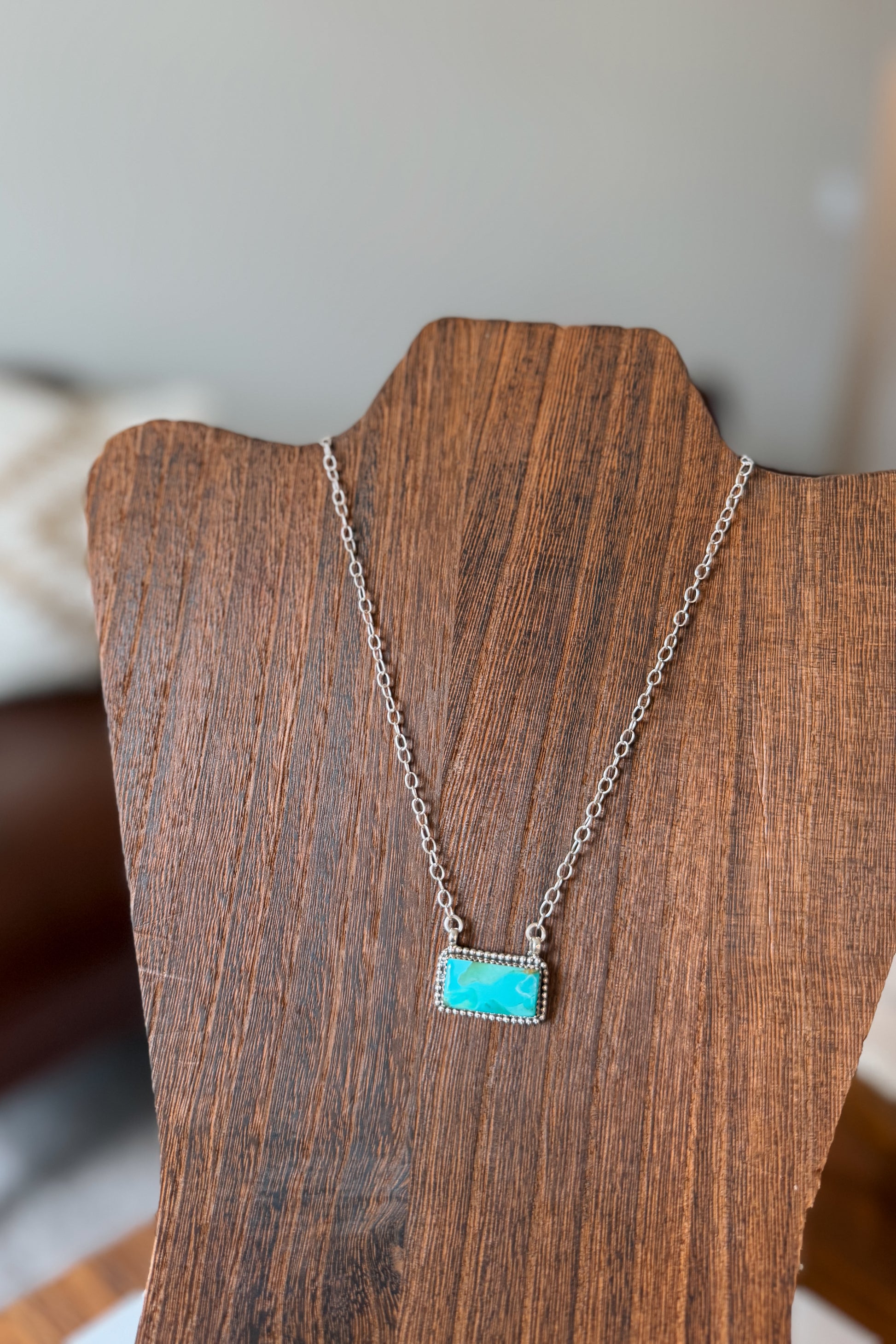 A silver chain necklace with a rectangular turquoise stone pendant, displayed on a dark wood necklace stand.