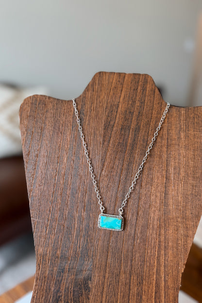 A silver chain necklace with a rectangular turquoise stone pendant, displayed on a dark wood necklace stand.