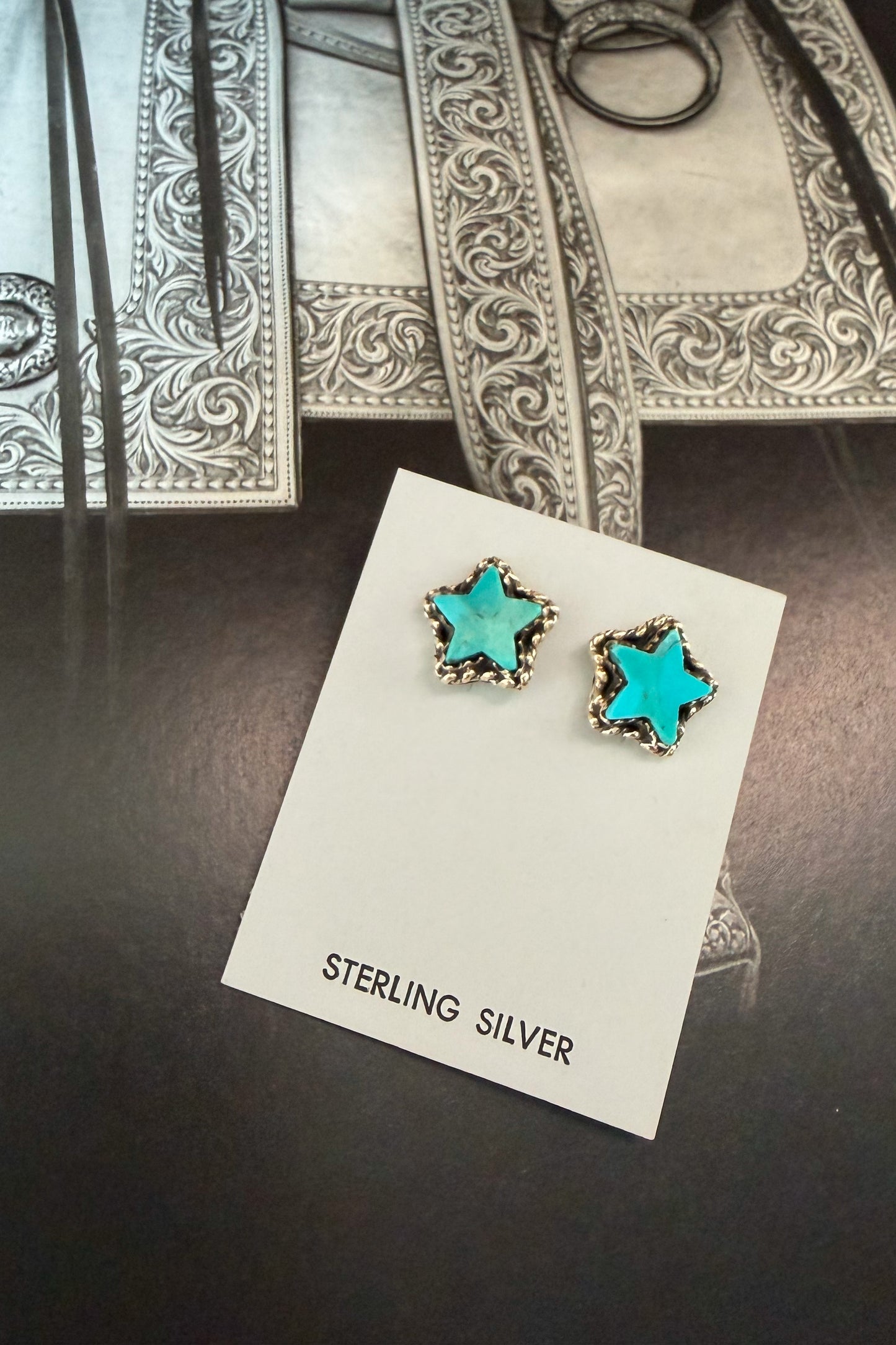 A pair of star-shaped stud earrings with turquoise blue centers and silver borders, displayed on a white card labeled 'STERLING SILVER' on a dark tabletop.