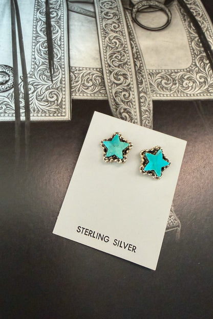 A pair of star-shaped stud earrings with turquoise blue centers and silver borders, displayed on a white card labeled 'STERLING SILVER' on a dark tabletop.