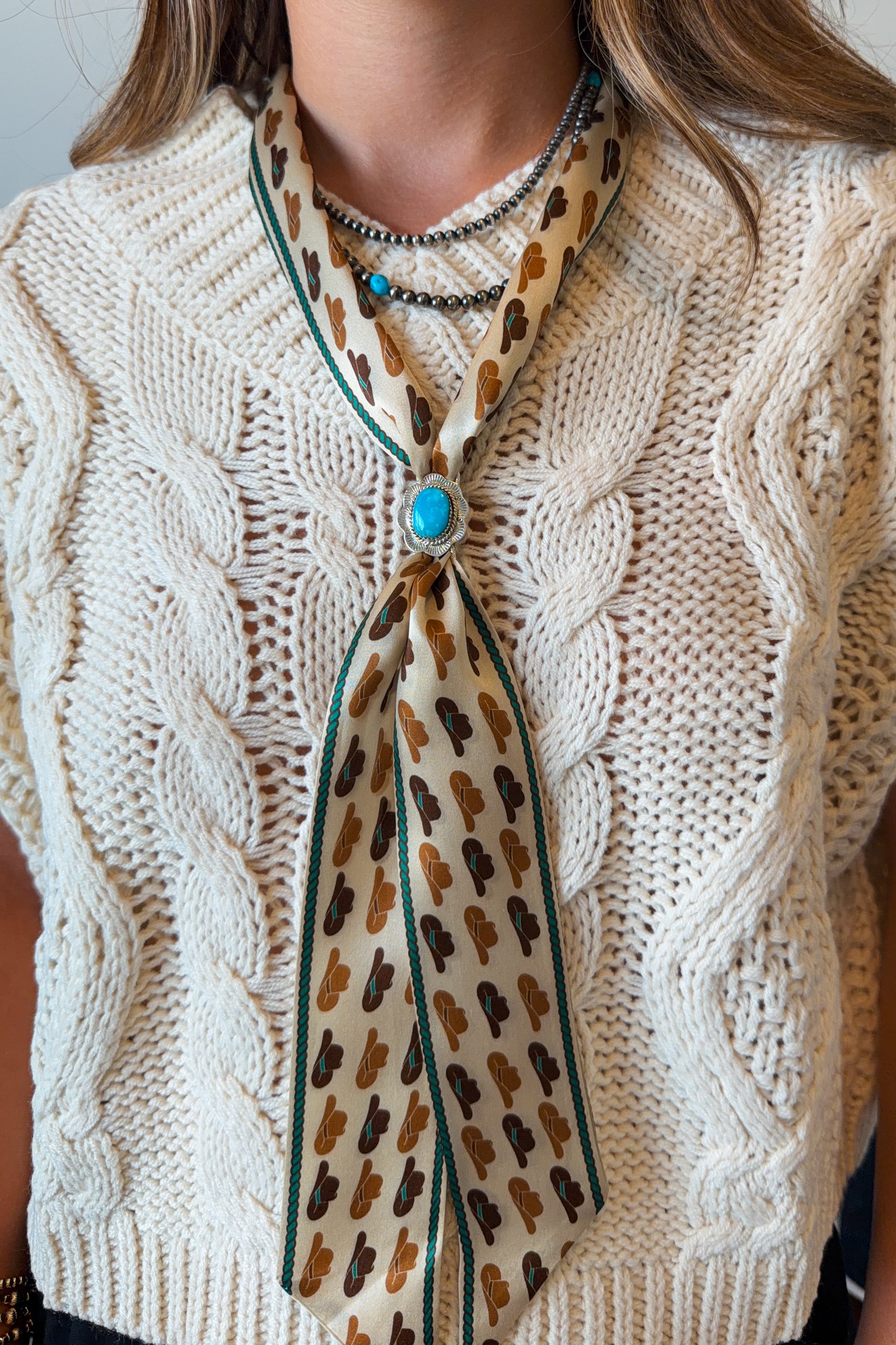 A close-up of a person wearing a cream cable-knit sweater vest over a white top, styled with a patterned scarf featuring a brown and tan print, dark green trim, and a turquoise stone accent.