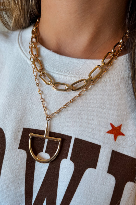 A close-up of a person wearing layered gold necklaces, including a chunky chain and a delicate chain with a D-ring pendant, over a white graphic sweatshirt.