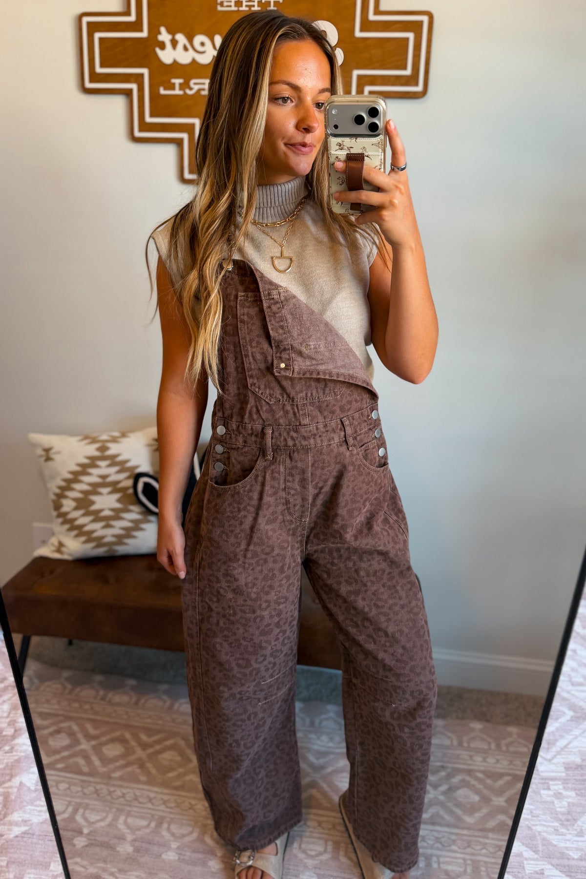 A woman takes a mirror selfie wearing brown leopard-print wide-leg overalls over a beige knit top.