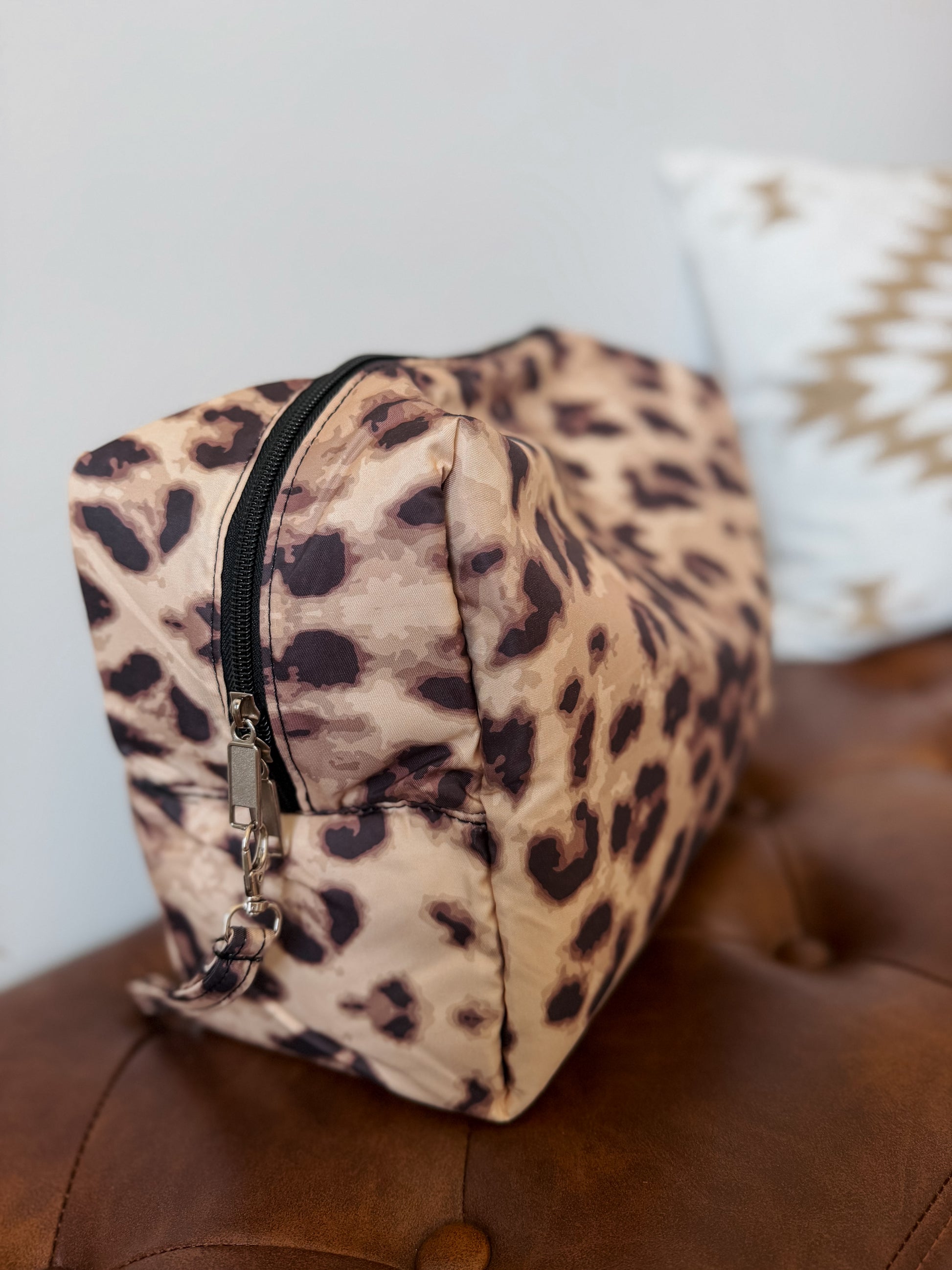 A leopard print cosmetic pouch with a black zipper, resting on a brown leather tufted surface with a white pillow in the background.