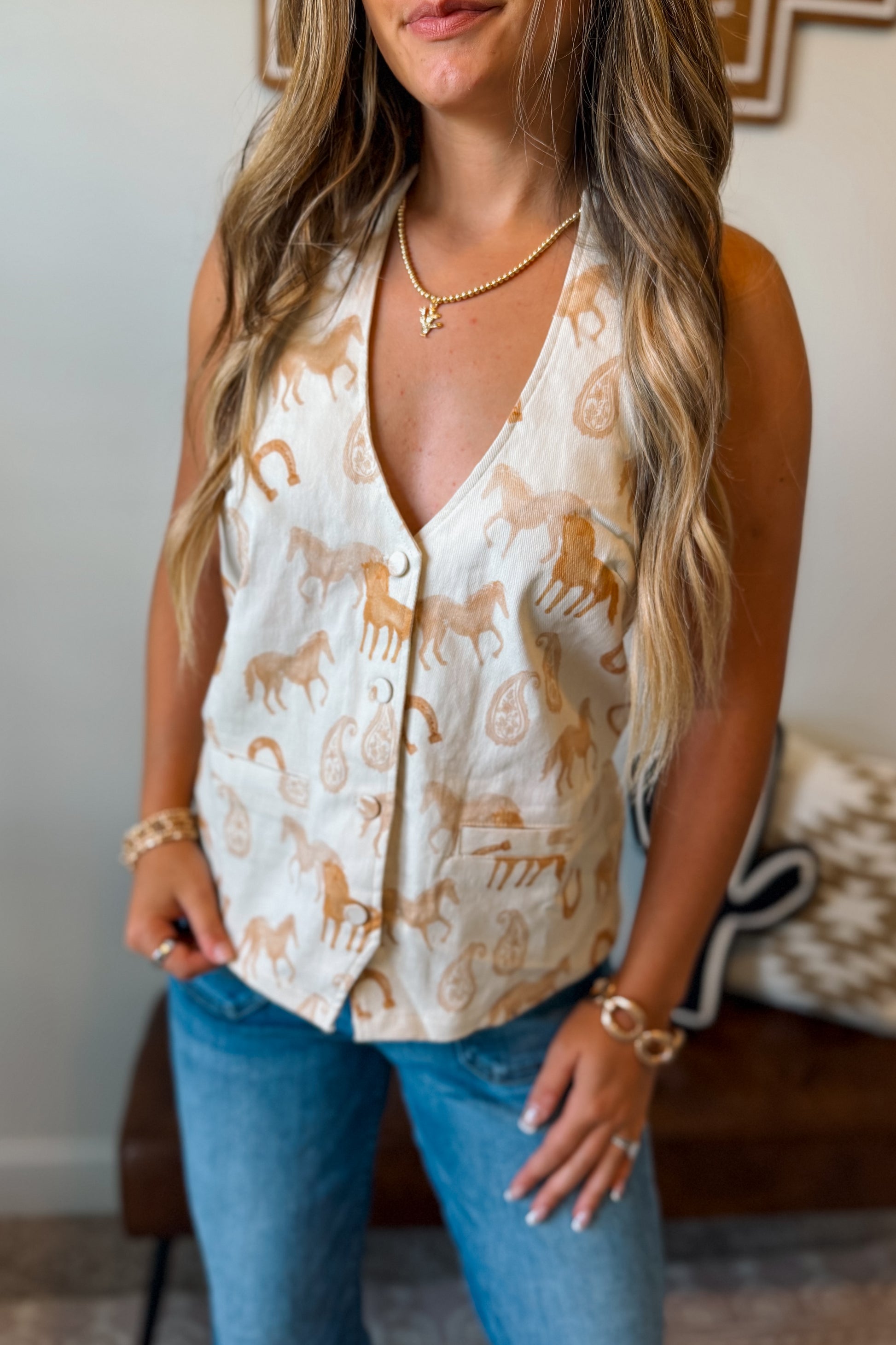 Close up front angle women wearing a sleeveless cream vest with a tan horse and paisley print, paired with blue jeans