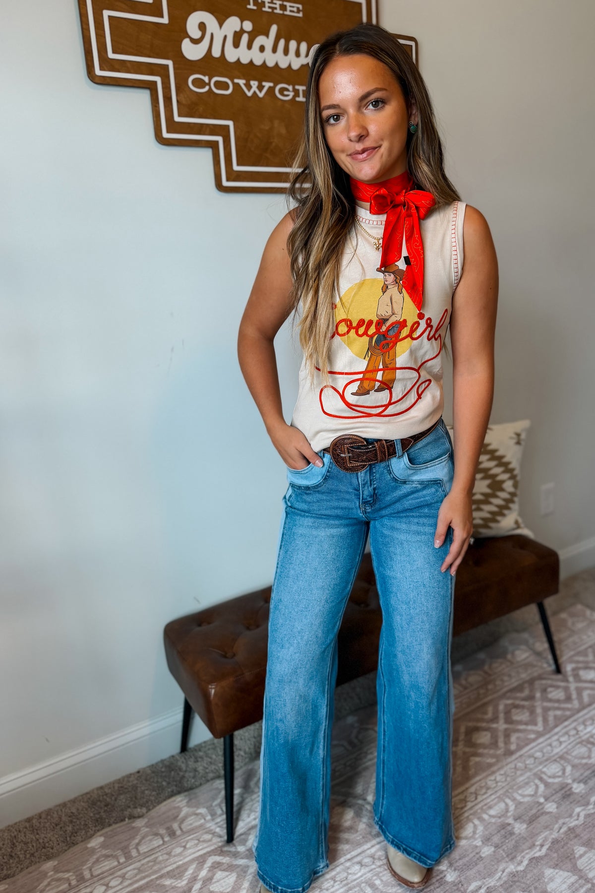 A woman modeling a sleeveless cream graphic top with a red neck scarf, blue flare jeans, and a brown belt.