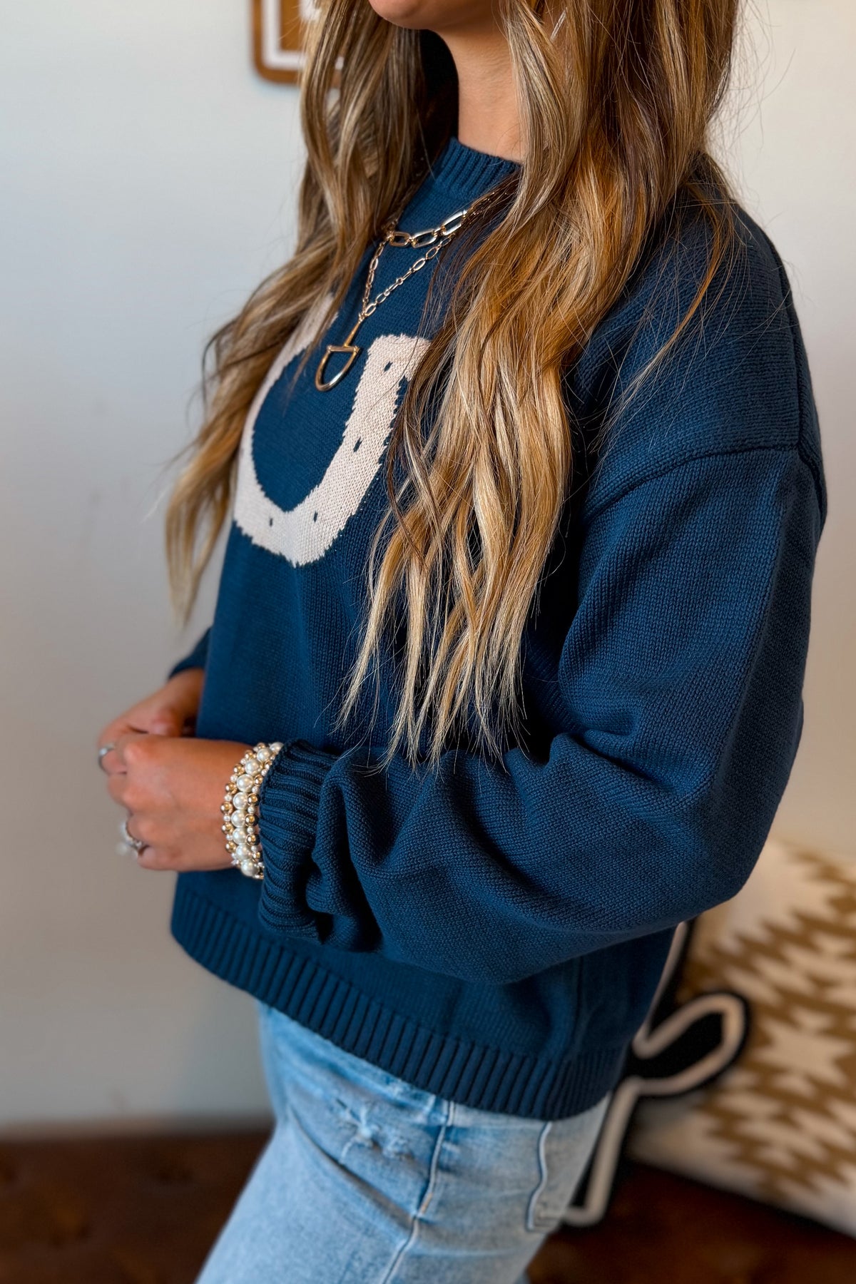 A woman wearing a navy blue knit sweater with a white horseshoe graphic on the chest, paired with light blue jeans and gold jewelry.