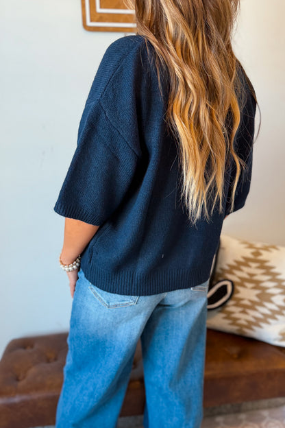 A woman shown from the back wearing an oversized dark navy knit sweater and light blue jeans, standing indoors.