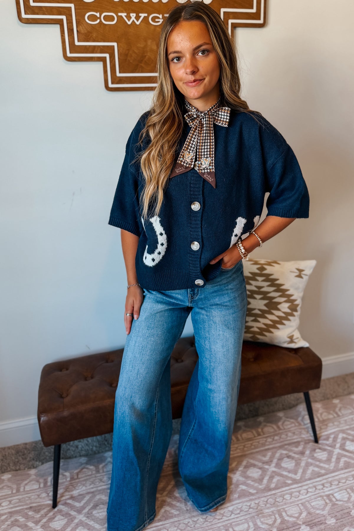A woman models a dark blue denim short-sleeve button-up cardigans with white horseshoes and a checkered necktie collar, paired with wide-leg blue jeans, standing in a styled indoor setting.
