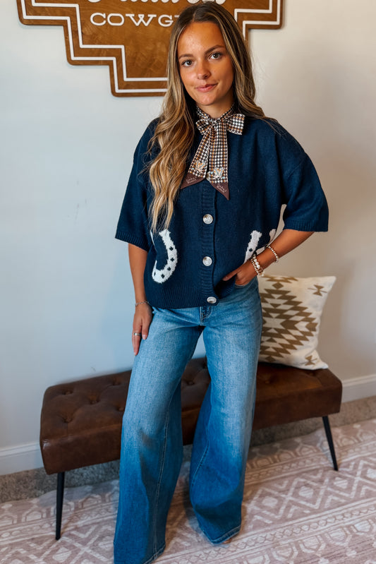 A woman models a dark blue denim short-sleeve button-up cardigans with white horseshoes and a checkered necktie collar, paired with wide-leg blue jeans, standing in a styled indoor setting.