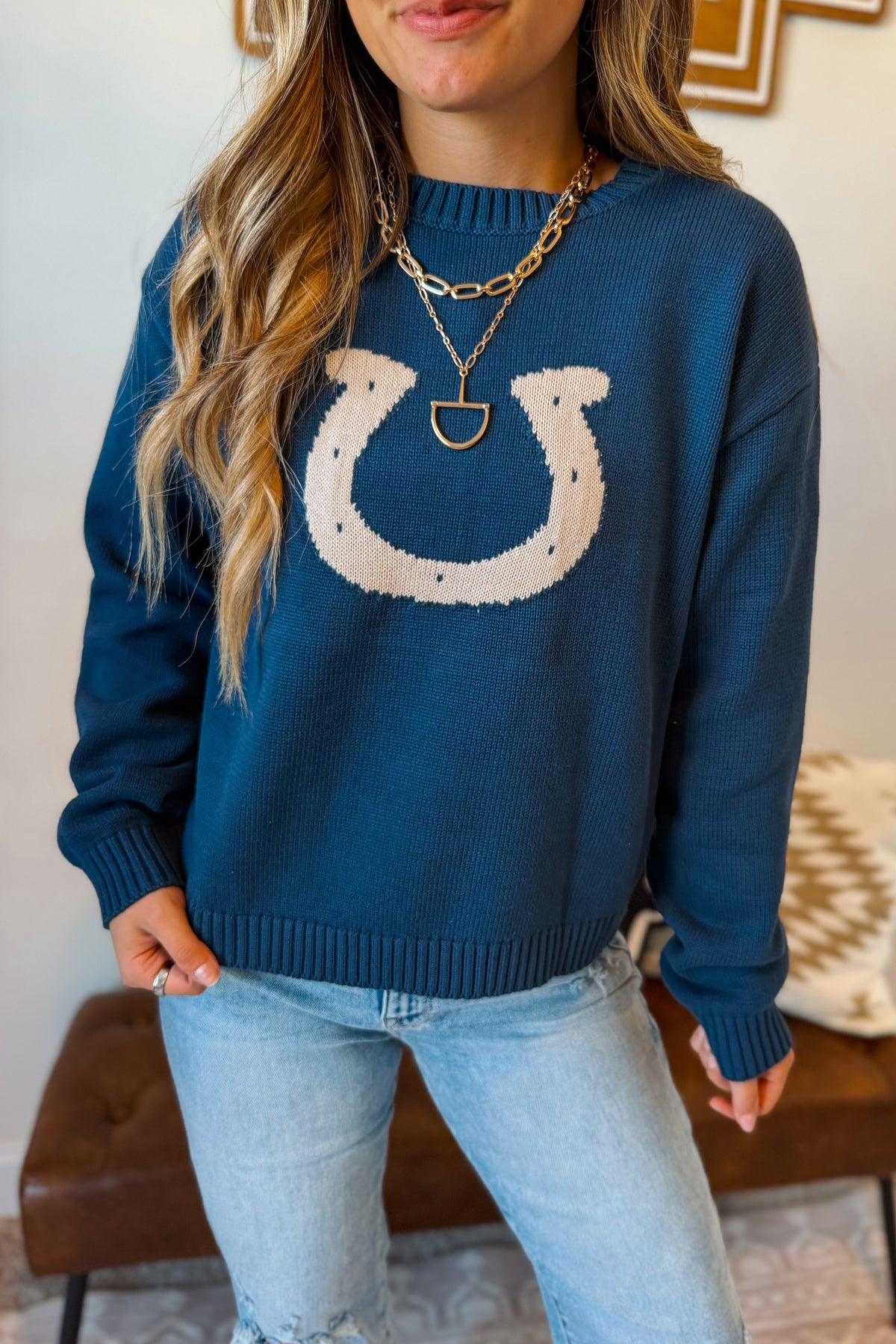 A woman wearing a navy blue knit sweater with a large white horseshoe graphic on the front, paired with light-wash jeans and layered gold necklaces.