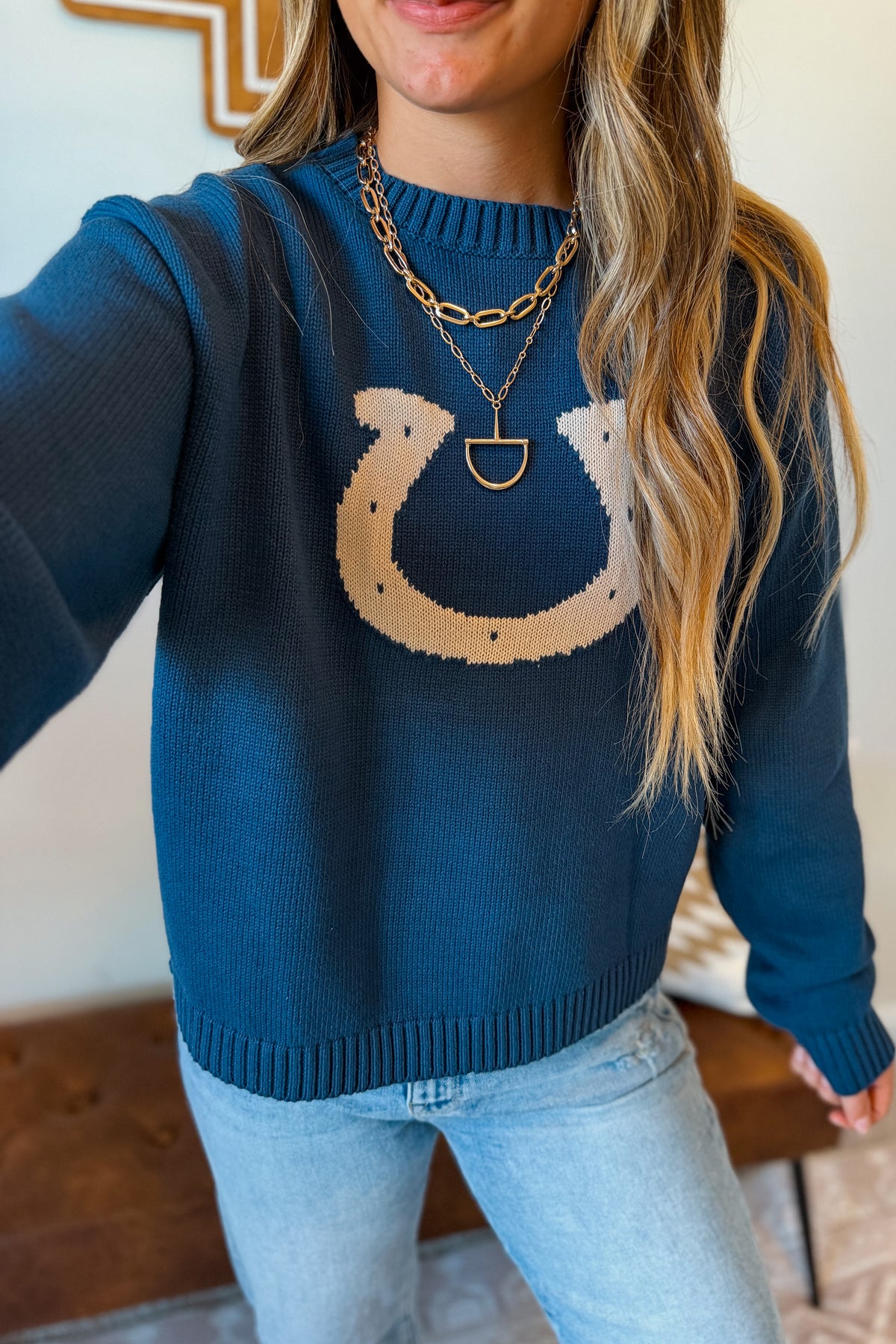 A woman wearing a navy blue knit sweater with a large beige horseshoe graphic on the chest, paired with light blue jeans and layered gold necklaces.