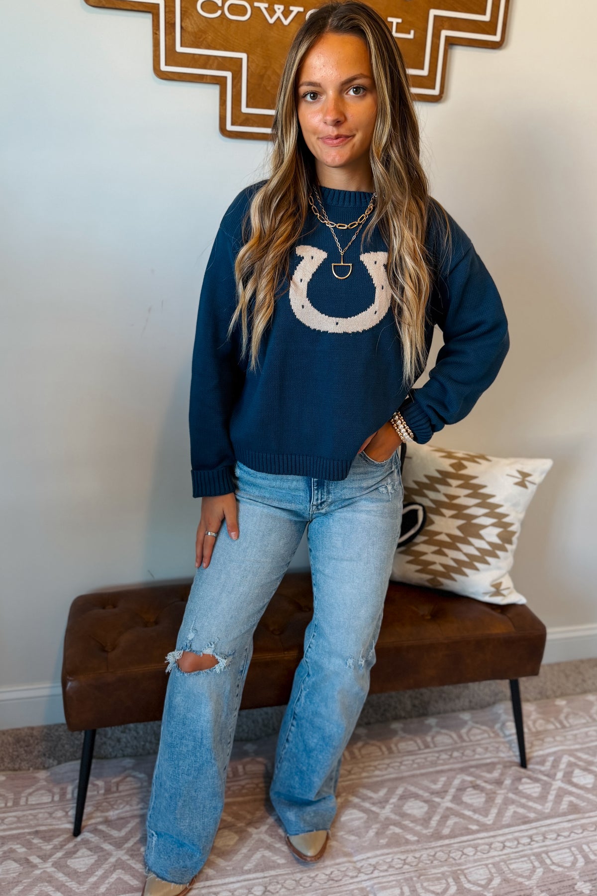 A woman wearing a navy blue crewneck sweater with a white horseshoe graphic on the chest, paired with light-wash distressed jeans