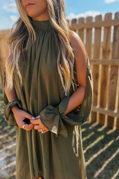 A woman wearing an olive green high-neck dress with cold-shoulder sleeves and tie details at the cuffs, standing outdoors in front of a wooden fence.