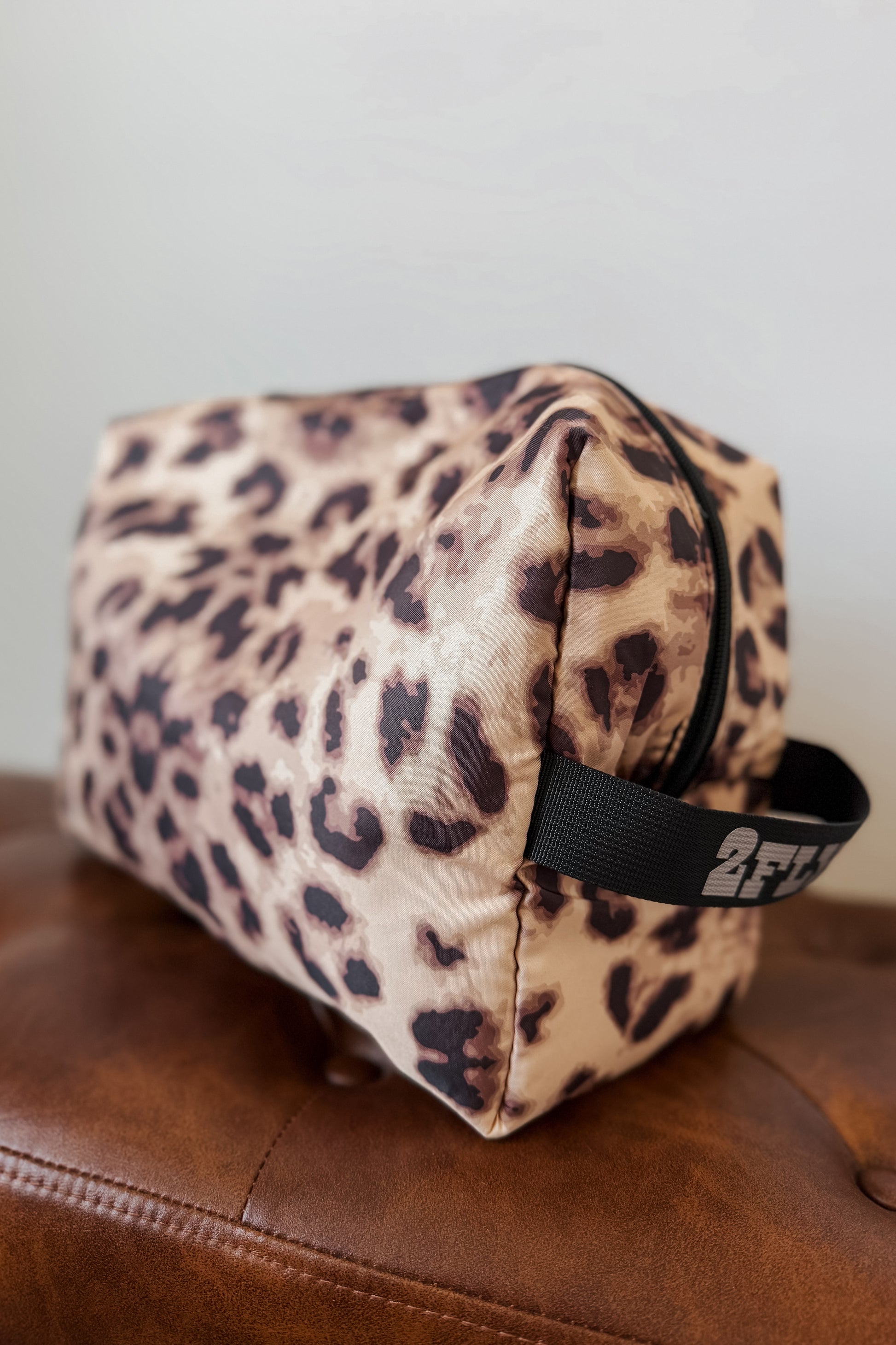 A leopard print cosmetic pouch with a black handle, resting on a brown leather surface