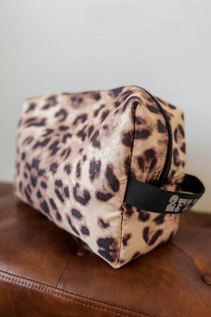 A leopard print cosmetic pouch with a black handle, resting on a brown leather surface
