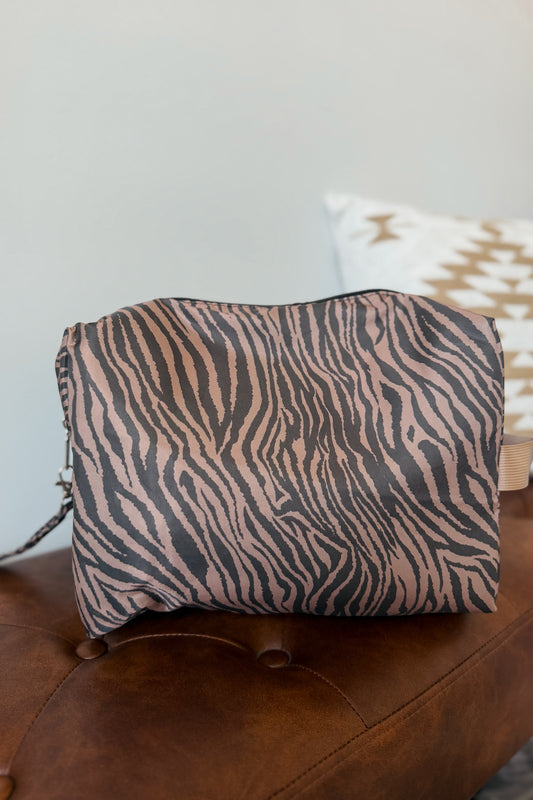 A zebra-print cosmetic pouch in dusty pink and black, resting on a brown leather bench
