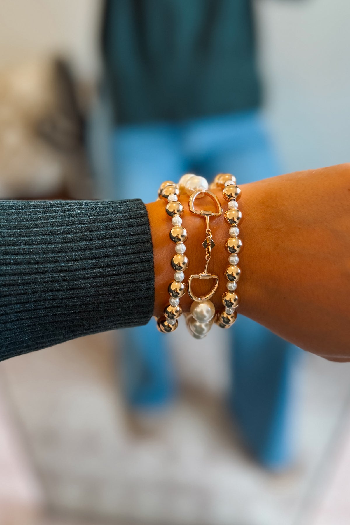 A close-up of a person's wrist wearing three stacked bracelets: gold and white bead strands, a gold horse bit charm bracelet, and a white textured pearl bracelet, styled with a dark ribbed knit sleeve.