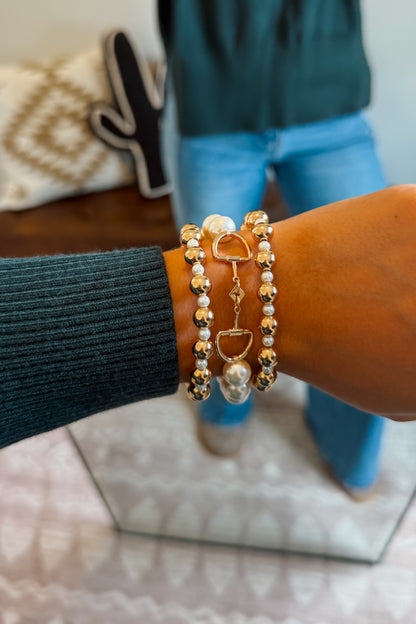 A close-up of a person's wrist wearing three beaded bracelets with gold-toned and white pearl-like beads, a small heart charm, and a snaffle bit-shaped detail.