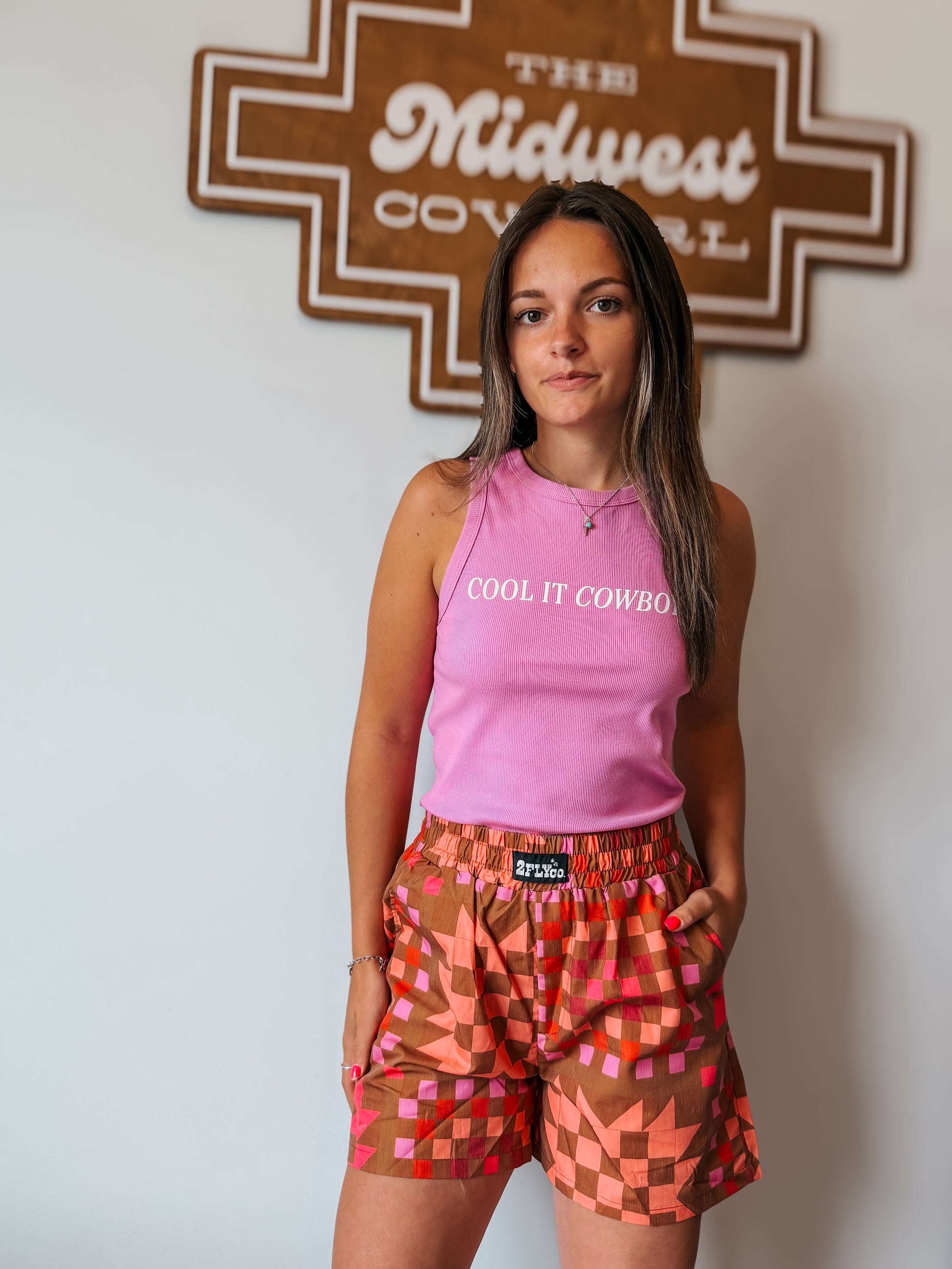 Woman wearing a pink tank top and patterned shorts standing in front of a sign that reads 'The Midwest Cowgirl'.