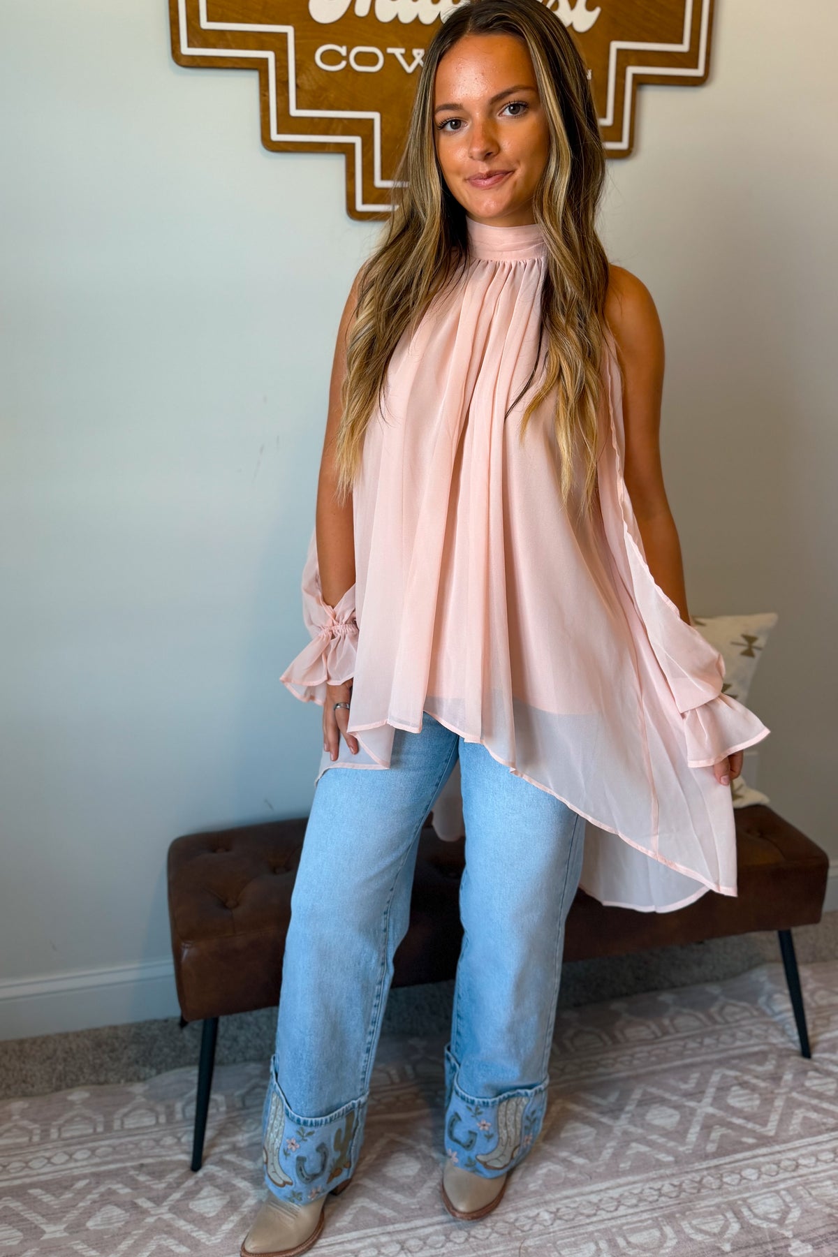A woman models a light pink sleeveless high-neck blouse with ruffled sleeves and an asymmetrical draped hem, paired with light blue jeans and tan boots